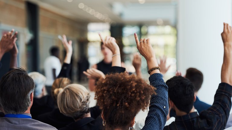 A group of sitting adults in a lecture, each with one of their hands up as if wanting to ask a question to the lecturer