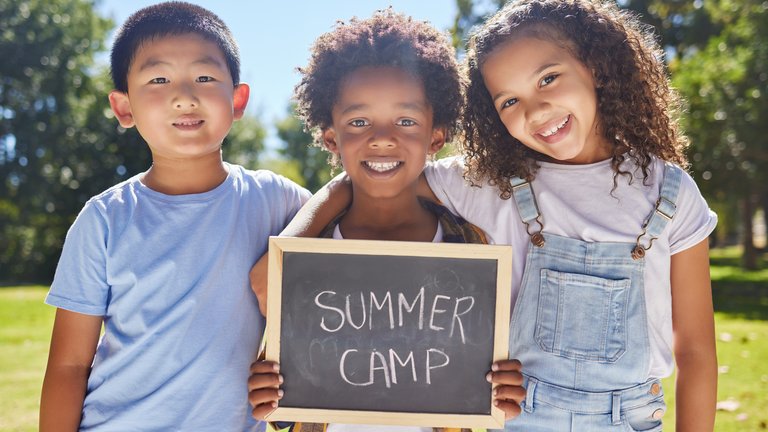 Three children in arms in a green field holding a small chalkboard that read's "Summer Camp"