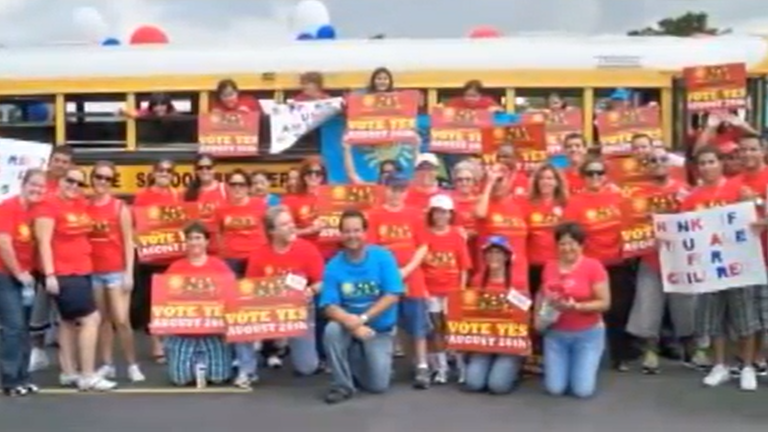 Group in red shirts holding 'Vote Yes' signs by a school bus.