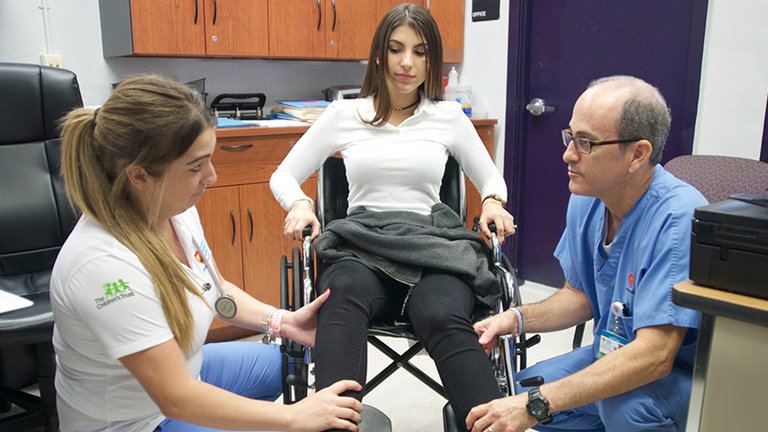 Patient in wheelchair being examined by two healthcare workers.