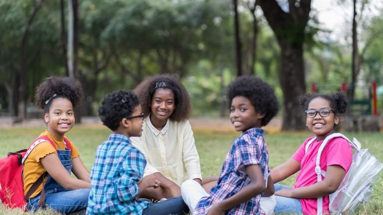 A group of smiling kids sitting in circle in grass with backpacks
