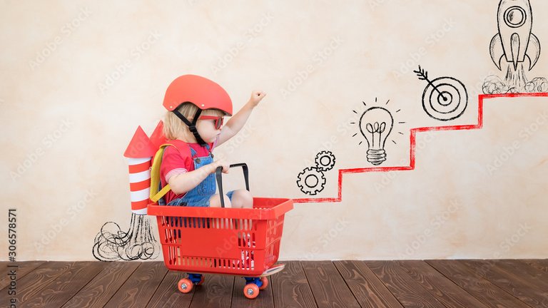 Child wearing a helmet and goggles, sitting in a shopping basket with a cardboard rocket on their back.