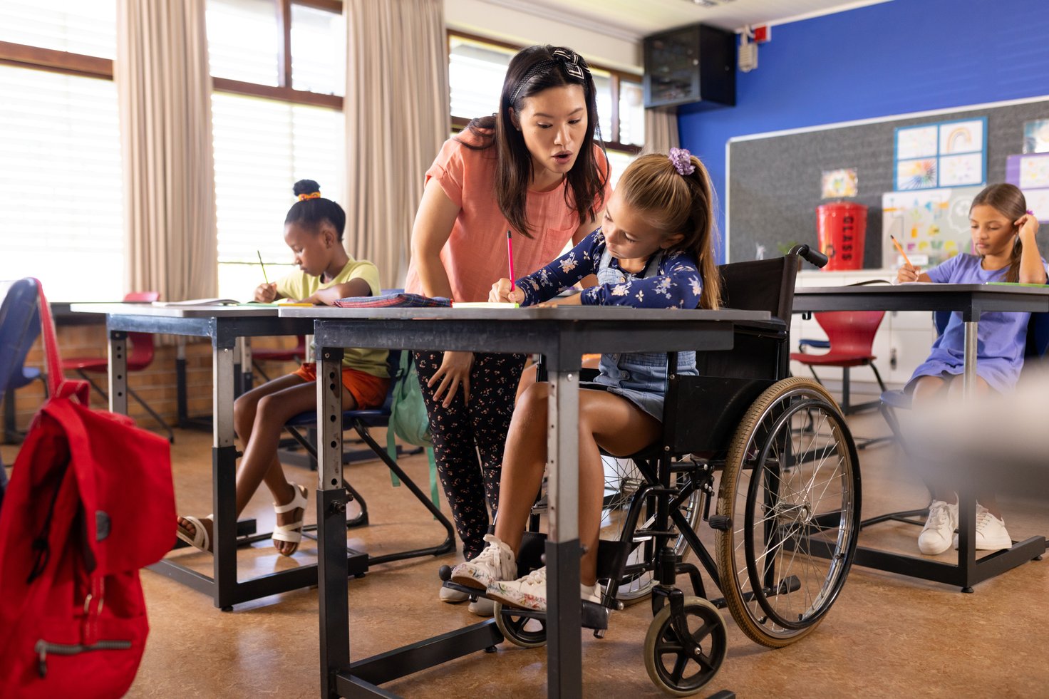 Teacher assisting a young girl in a wheelchair with her classwork in an inclusive classroom setting.