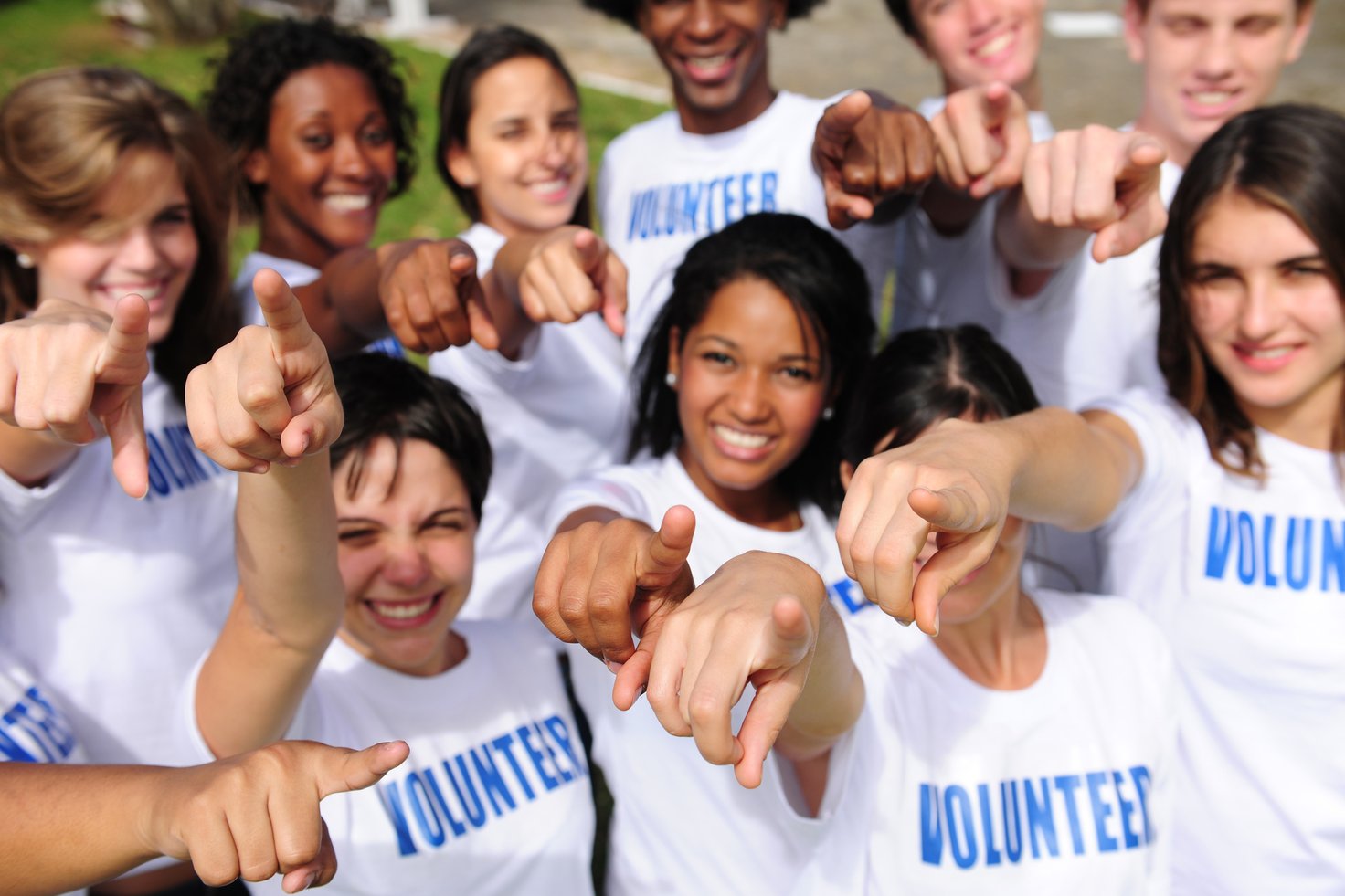 Group of diverse volunteers smiling and pointing towards the camera, wearing matching shirts that say 'VOLUNTEER'