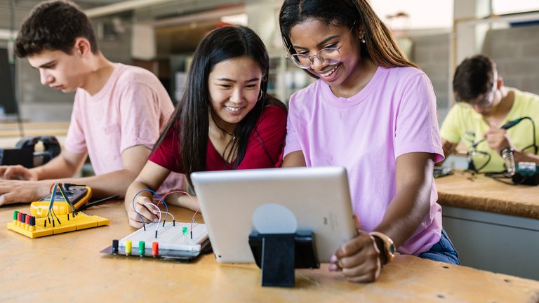Two students collaborating on a tablet in a classroom, working on an electronics project with circuits and breadboards.