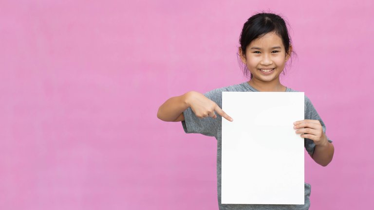 A young girl holding a blank posterboard while standing in front of a pink wall