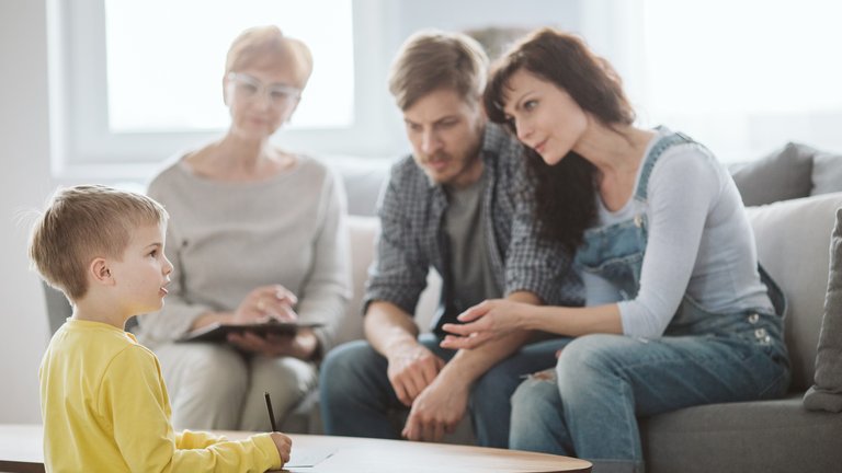 Three adults sitting on a couch, one therapist, two parents, and a child on the other end of a coffee table presenting something