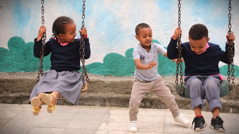 three kids playing on swings, one kid is pushing another kid on the swing.