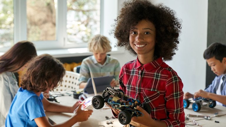A teenager holding a remote controlled car, smiling in a classroom with classmates.