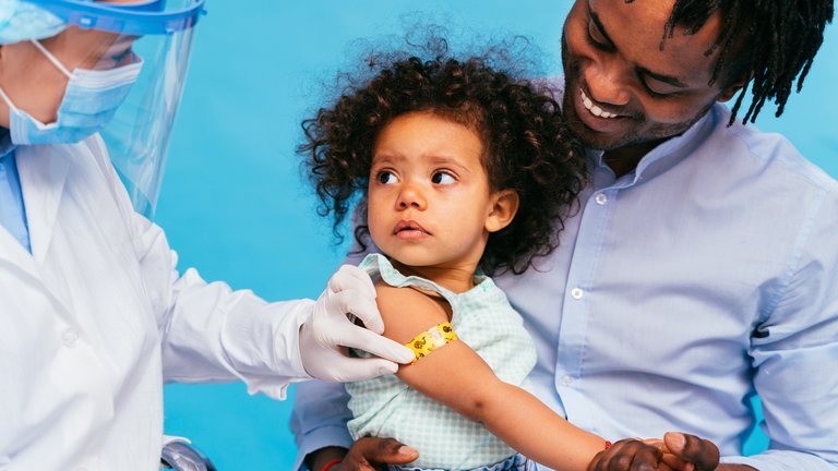 Health worker applying a colorful bandage to a little girl's arm being held by her father.