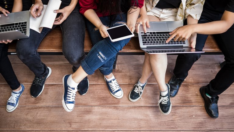 A group of people sitting in a wooden bench, holding and pointing at tech gadgets and a notebook.