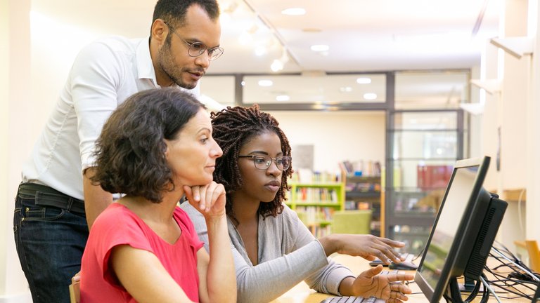 Three adults collaborating at a computer workstation in a bright, modern classroom or library setting.